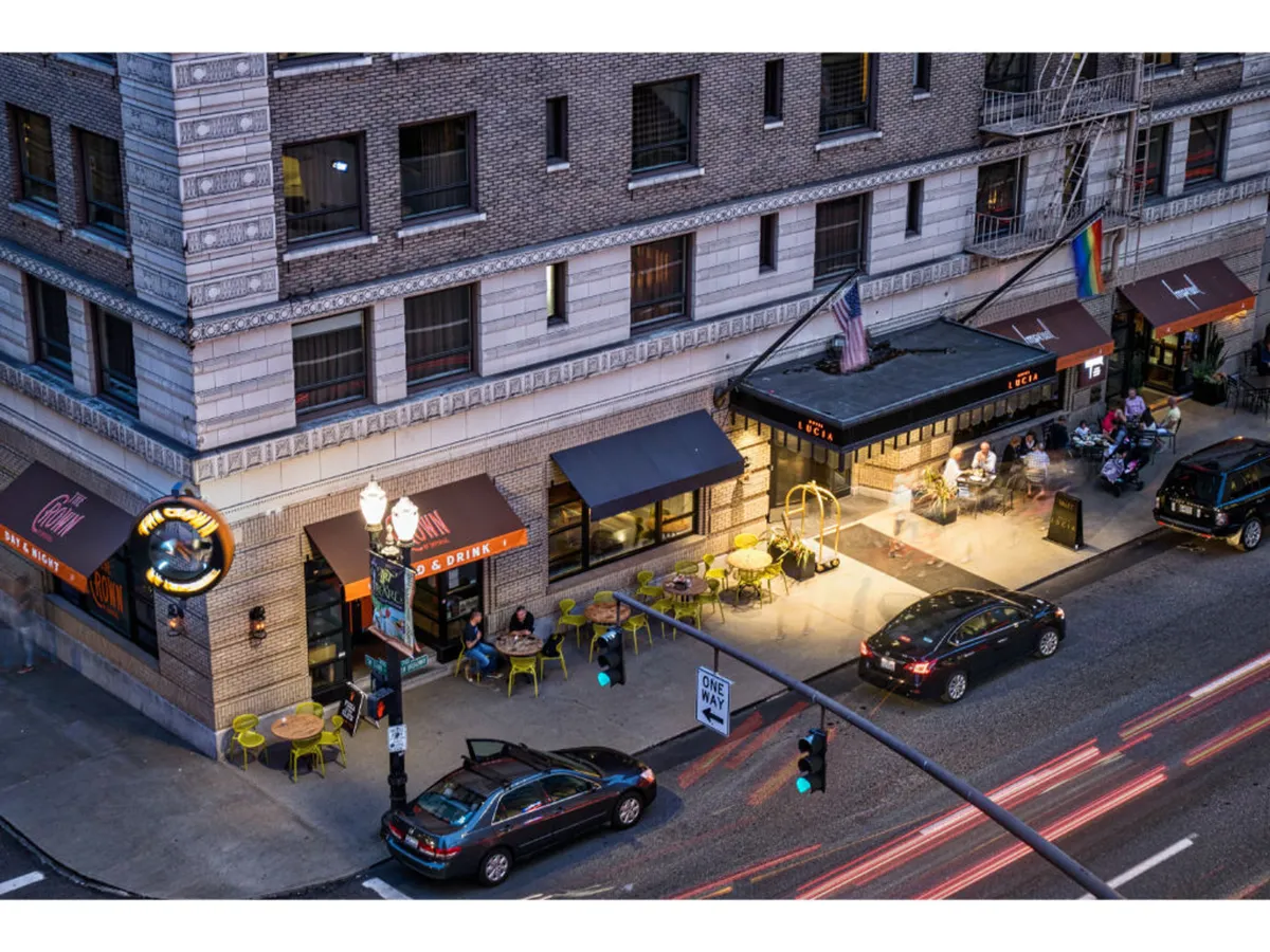 An inviting restaurant patio with outdoor seating, street lights, and cars passing on the road, evoking a vibrant urban atmosphere. The scene suggests a lively city environment ideal for socializing, relaxing, and enjoying local food and drinks.
