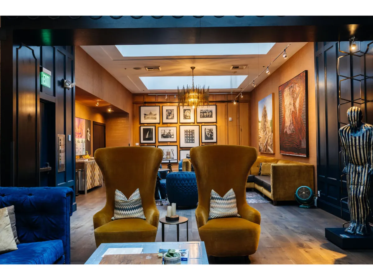A vibrant interior lounge with two large yellow armchairs, a blue couch, and striking framed photographs and artistic wall paintings. A metal sculpture adds a creative touch, and the seating area invites relaxation and conversation amid curated art displays.