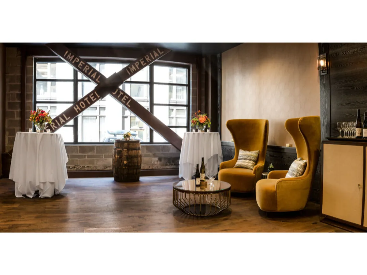 A cozy lounge area featuring two yellow armchairs, small round tables, decorative floral arrangements, and a barrel. A large window with signage allows natural light to flow in, subtly hinting at the hotel's connection to the city outside.