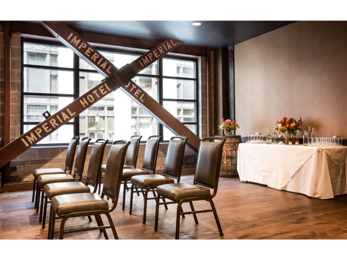 A sophisticated gathering space with a decorative crossbeam inscribed 'IMPERIAL HOTEL,' carefully arranged chairs, a table set with floral centerpieces and glassware, and an overall formal yet welcoming atmosphere. Ideal for meetings, workshops, or social events.