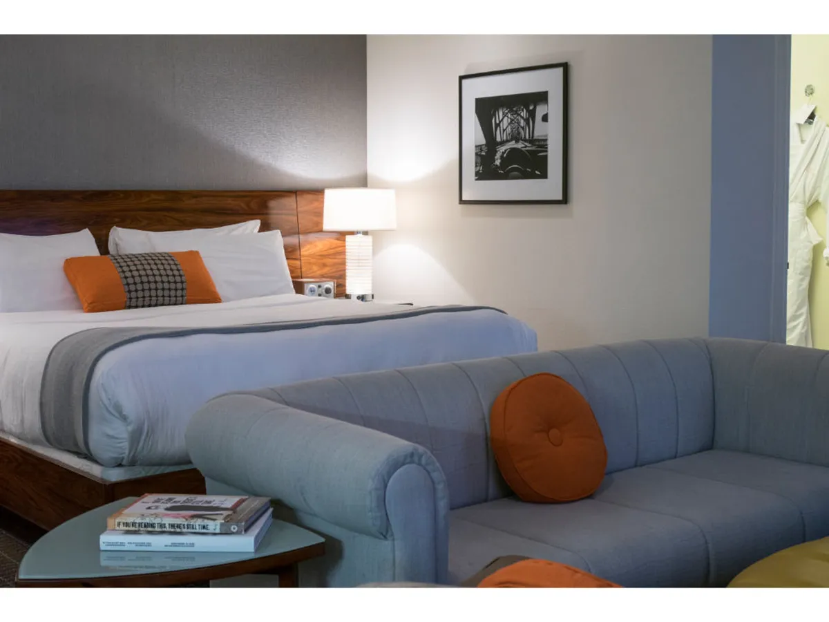 The image shows a stylish hotel room interior featuring a plush bed adorned with decorative pillows, a chic gray couch with a striking orange accent, and a table topped with inviting books. The cozy atmosphere suggests a space ideal for sipping coffee in the morning, unwinding with a good read, or connecting with friends after a day exploring the city.