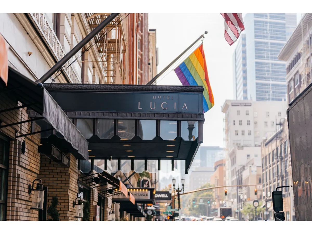 An exterior shot of Hotel Lucia featuring its sign, a Rainbow flag, and American flag. The street is lively, suggesting a vibrant, inclusive urban setting just steps from the hotel’s entrance. The image evokes the hotel’s central location and accessibility to city adventures.