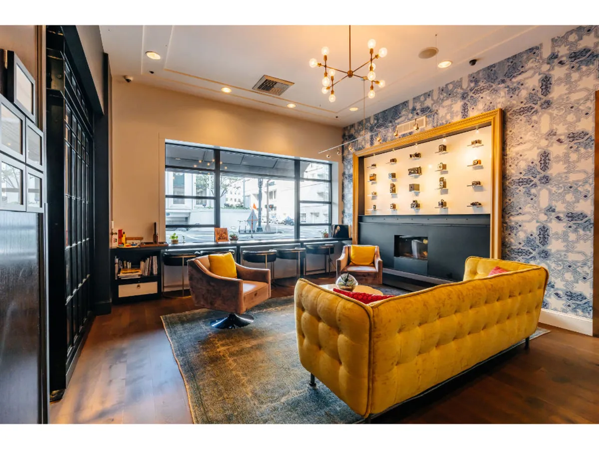 A welcoming lounge area featuring a vibrant yellow tufted sofa, charming armchairs, decorative wall shelves, and large windows that let in natural light. The space is suitable for both casual relaxation and productive work, with guests seen reading and working on laptops.