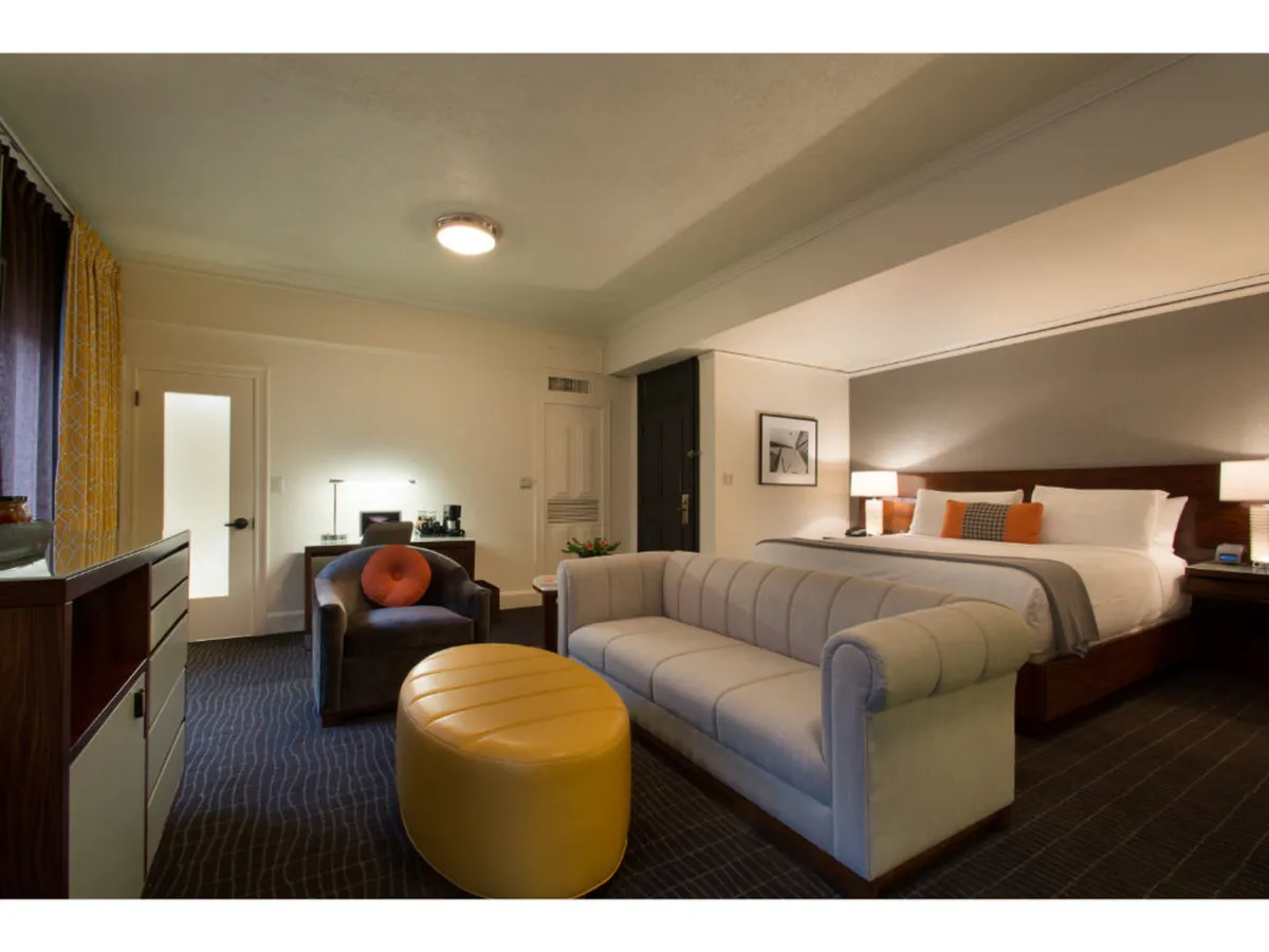 A wide view of the suite’s living and working area, including a plush sofa, ottoman, bed, and a dedicated desk. The arrangement highlights the room’s comfort and practicality, with natural light illuminating both restful and productive spaces.