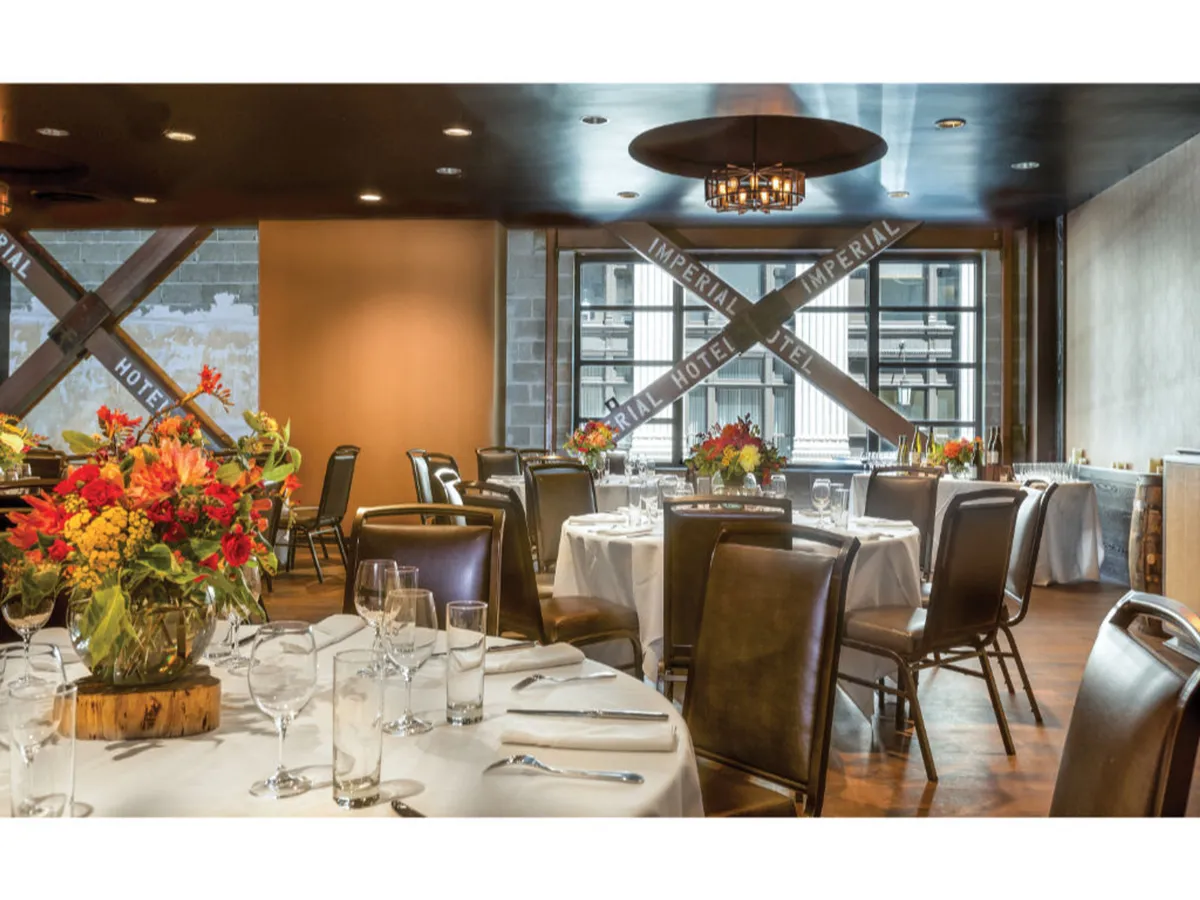 A sophisticated dining area with circular tables adorned with decorative floral arrangements. Large windows offer expansive city views, while a chandelier adds an elegant touch. The space is bright, inviting, and visually connects the hotel to the vibrant city outside.