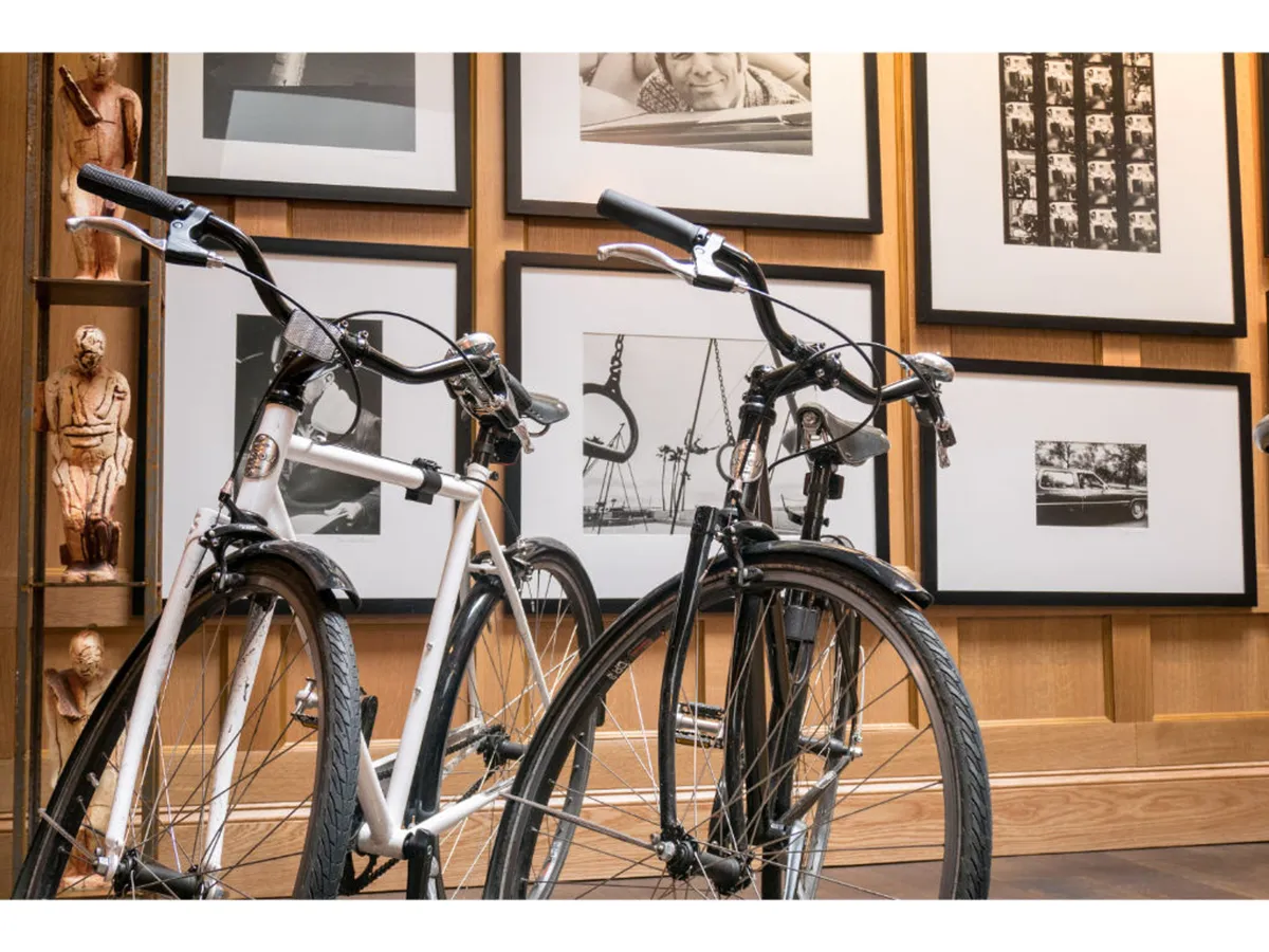 This image showcases two bicycles (one white, one black) leaning against a wooden wall adorned with black and white framed photographs and other artwork. The setting suggests a creative common area that encourages exploration and active lifestyles, with a nod to local art and culture.