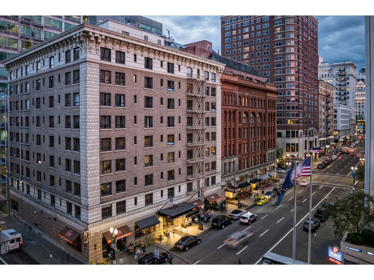 An exterior street scene featuring historic buildings illuminated at dusk, bustling street activity, and city lights. Sidewalk cafes and a lively pedestrian presence convey the energetic atmosphere of downtown Portland.