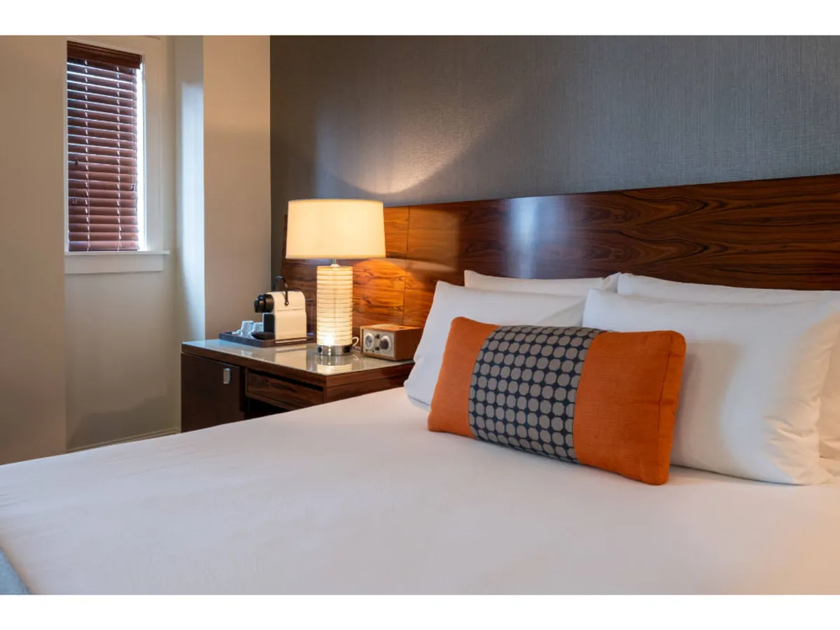 A serene bedroom with a plush bed adorned with decorative pillows, a table lamp on a nightstand, and soft natural light creating a relaxing ambiance. It highlights a tranquil retreat for rest and rejuvenation, ideal for either leisure or business travelers.