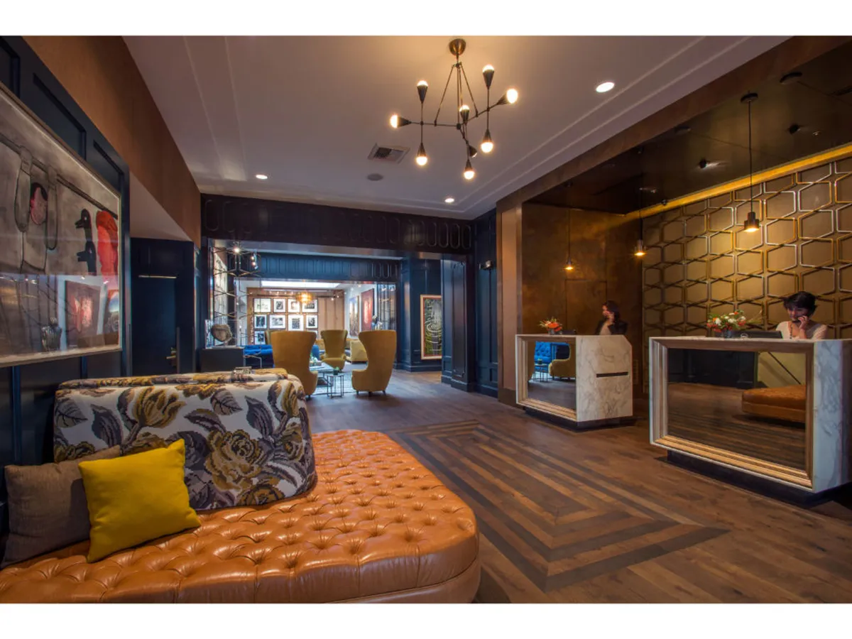 A contemporary and welcoming lobby area featuring a tufted orange sofa, vibrant armchairs, artistic wall decor, and unique reception desks. Guests are seen enjoying the space, checking in, and relaxing among the decor.