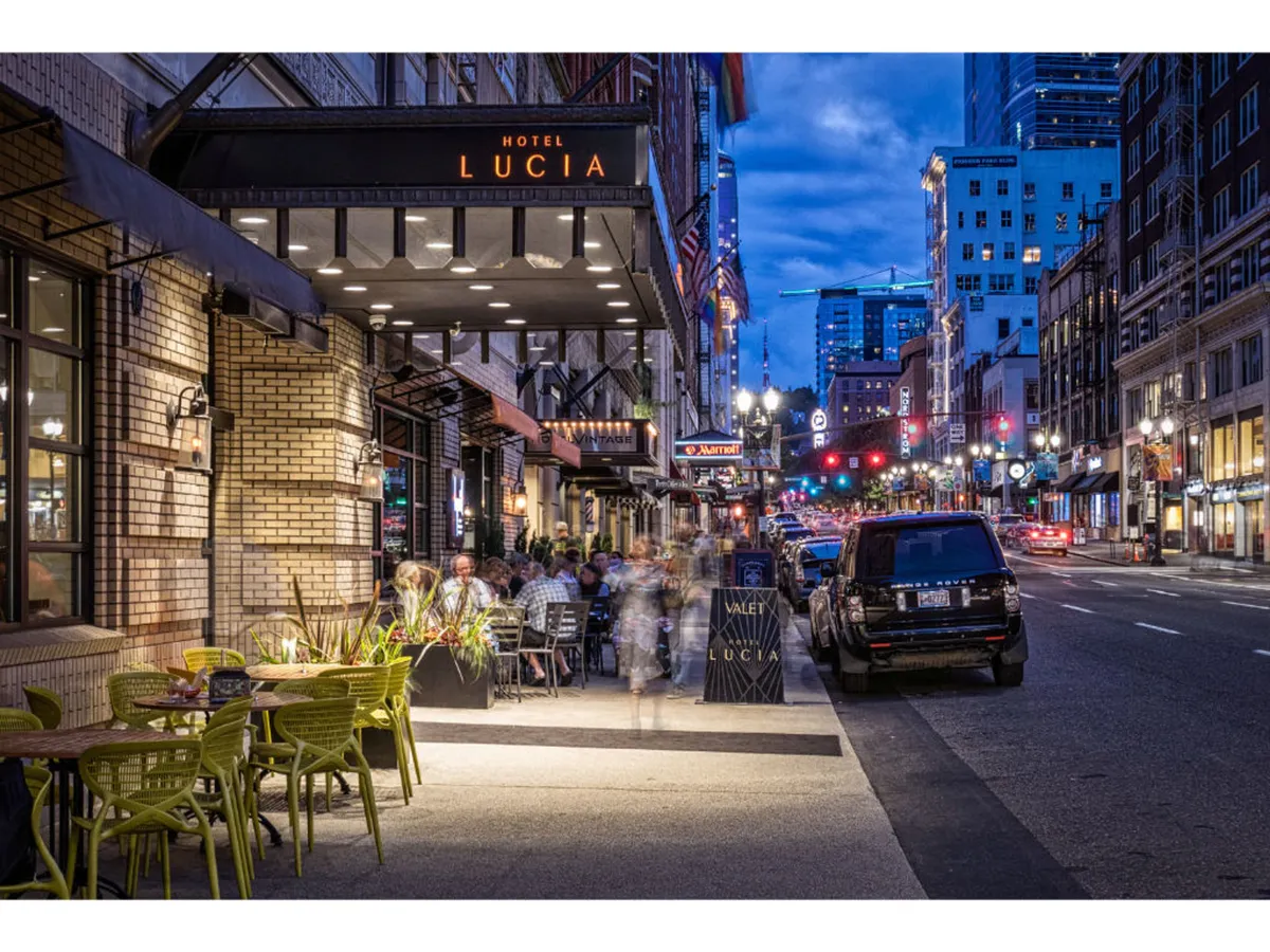 A lively nighttime exterior view of Hotel Lucia, with the hotel entrance, restaurant seating spilling out onto the sidewalk, people dining outdoors, illuminated street lights, and a glimpse of the city skyline in the background. The setting is abuzz with city energy and social activity.