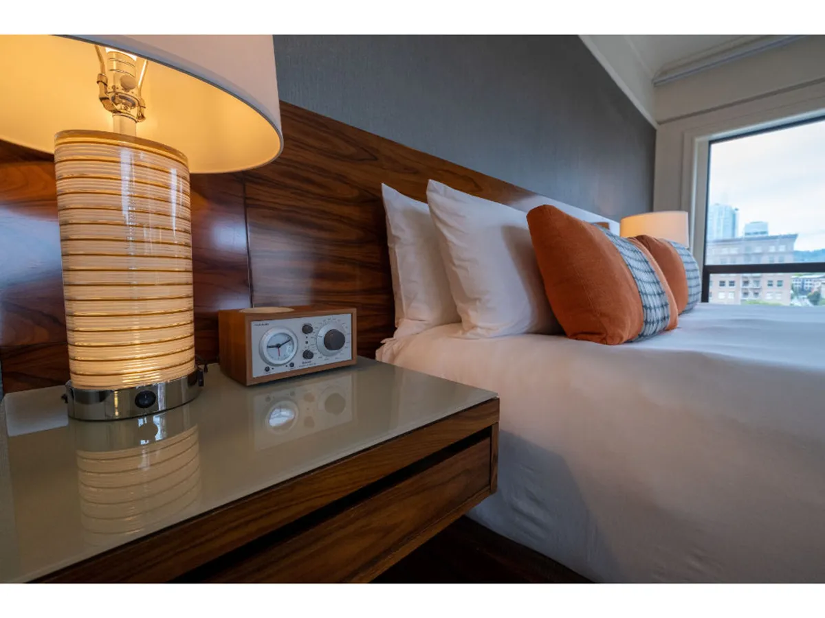 A serene scene of a bed with decorative pillows, a bedside lamp, and a nightstand, basking in soft natural light. The inviting setup hints at restful mornings and the hotel’s attention to guest comfort, ideal for those traveling with pets who appreciate a tranquil retreat.